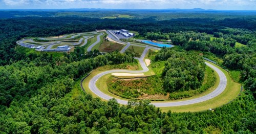 Aerial view of a winding racetrack surrounded by lush greenery and hills under a clear blue sky.