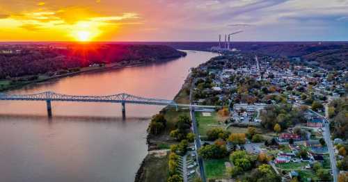 Aerial view of a river at sunset, with a bridge, a small town, and power plants in the background.