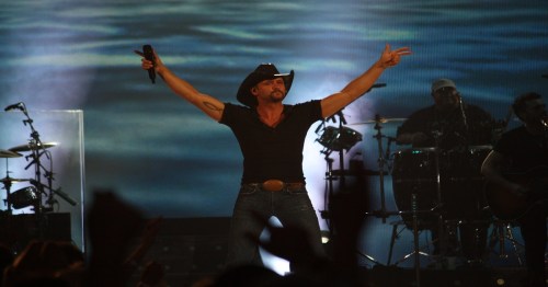 A male country singer in a cowboy hat performs energetically on stage, with band members and lights in the background.