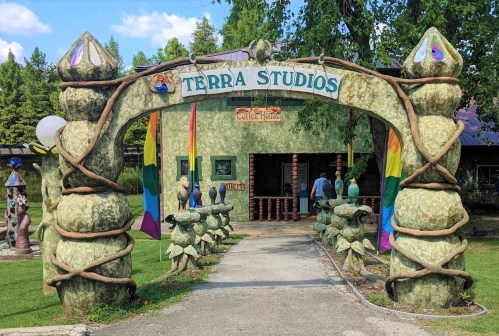 Colorful entrance archway to Terra Studios, featuring sculptures and rainbow flags, surrounded by greenery.