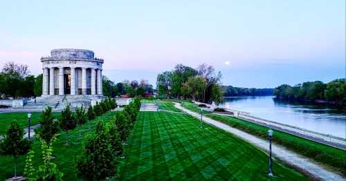 A scenic view of a monument by a river at dusk, with trees and a full moon in the sky.