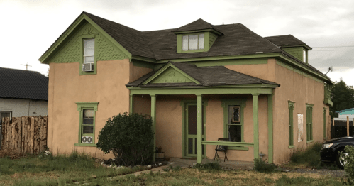 A two-story house with a peach exterior and green trim, featuring a front porch and overgrown yard.