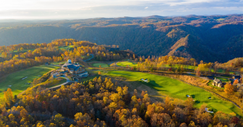 Aerial view of a golf course surrounded by autumn foliage and rolling hills under a clear sky.