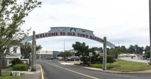 Sign reading "Welcome to Old Town Bandon" at the entrance of a small coastal town, with trees and buildings in the background.