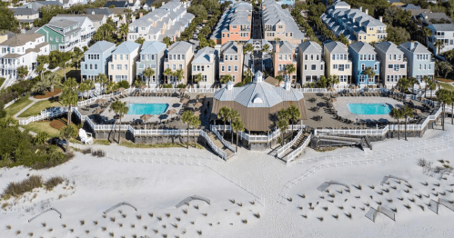 Aerial view of colorful beach houses with pools, surrounded by palm trees and sandy beach.