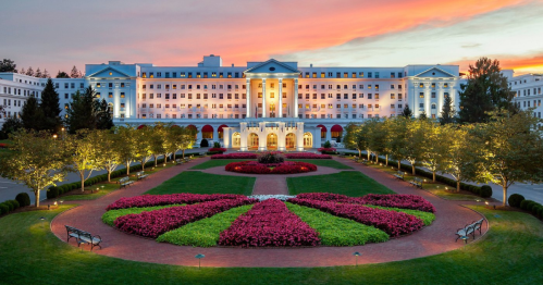 A grand hotel with a colorful flower garden at sunset, surrounded by trees and illuminated by soft lights.