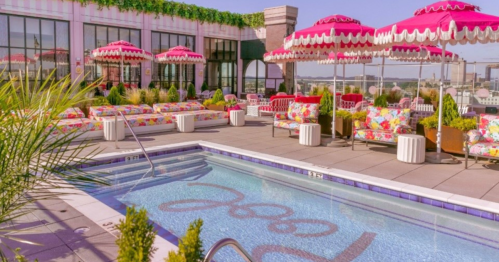 Rooftop pool area with colorful umbrellas, lounge chairs, and vibrant floral decor against a city skyline backdrop.