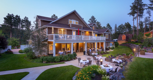 A charming two-story house with a porch, surrounded by trees and gardens, featuring outdoor dining tables.