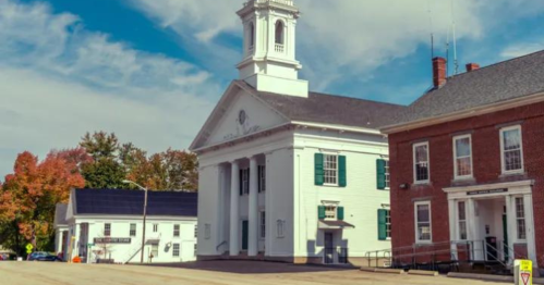 Historic white building with a clock tower, surrounded by trees and a red brick structure, under a blue sky.
