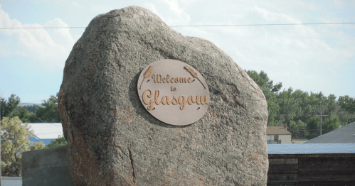 A large rock with a metal plaque reading "Welcome to Glasgow" against a blue sky and trees in the background.