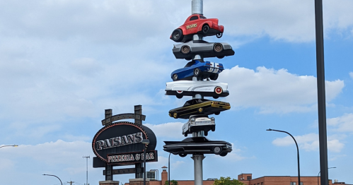 A pole topped with stacked toy cars next to a sign for Paisano's Pizzeria & Bar under a blue sky.