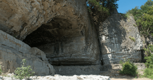 A rocky cliff with a large overhang, surrounded by greenery and blue sky. Some graffiti is visible on the rock surface.