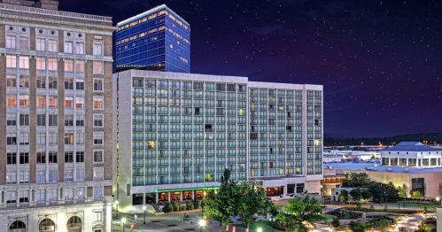 A modern hotel building at night, illuminated against a starry sky, surrounded by city lights and greenery.