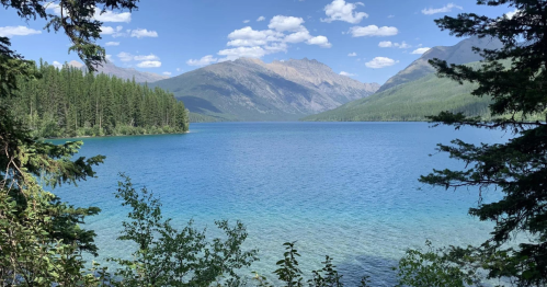 A serene lake surrounded by mountains and lush greenery under a clear blue sky with scattered clouds.