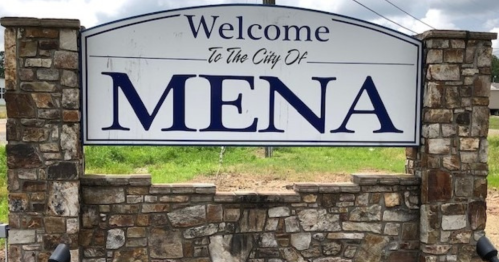 Welcome sign for the city of Mena, featuring a stone base and blue lettering against a cloudy sky.