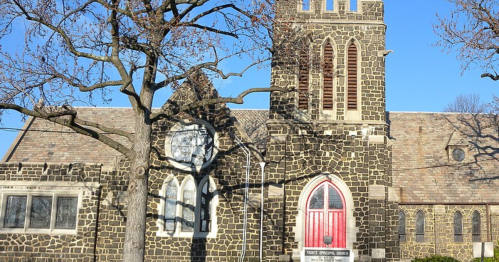 A stone church with a tall tower and red door, surrounded by bare trees against a clear blue sky.