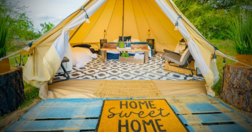 A cozy glamping tent with a welcome mat, wooden furniture, and a patterned rug, surrounded by nature.