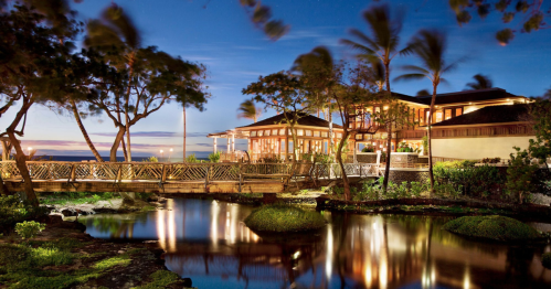 A serene waterfront view of a restaurant at dusk, surrounded by palm trees and reflecting in the calm water.