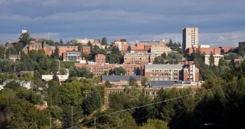 A panoramic view of a university campus surrounded by trees and buildings under a blue sky with scattered clouds.