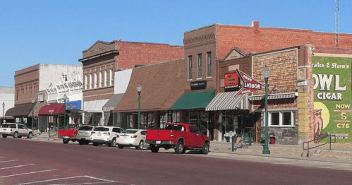 A row of historic buildings with shops and a liquor store, featuring brick facades and parked cars on a sunny day.