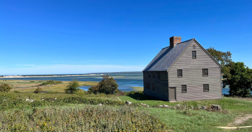 A historic wooden house beside a calm waterway, surrounded by green fields and a clear blue sky.