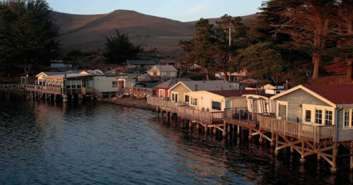 A serene waterfront scene with wooden houses on stilts, surrounded by trees and mountains at sunset.