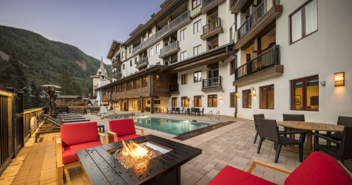 A modern hotel with a pool, outdoor seating, and a fire pit, set against a mountain backdrop at dusk.
