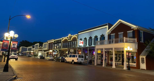 A charming street scene at dusk, featuring historic buildings adorned with lights and parked cars along the road.