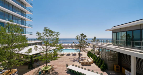 A sunny outdoor pool area with lounge chairs, trees, and ocean views, surrounded by modern buildings.