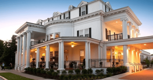 A grand white building with columns, large windows, and a welcoming porch, illuminated at dusk.