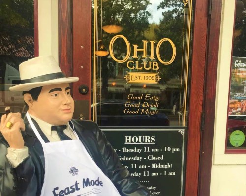 A statue of a man in a suit and hat stands outside the Ohio Club, showcasing its hours and offerings.