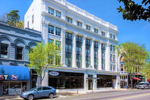 Historic building with columns and large windows, surrounded by trees and shops on a sunny day.