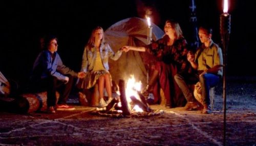 A group of four people sitting around a campfire at night, holding hands and engaged in a ritual or storytelling.