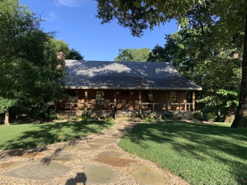 A charming wooden house with a stone pathway, surrounded by lush greenery and trees under a clear blue sky.