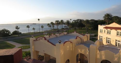 View of a waterfront with palm trees, a park, and a building in the foreground during sunset.
