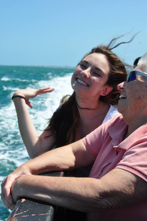 Two women enjoy a boat ride, smiling and laughing, with wind in their hair and ocean waves in the background.