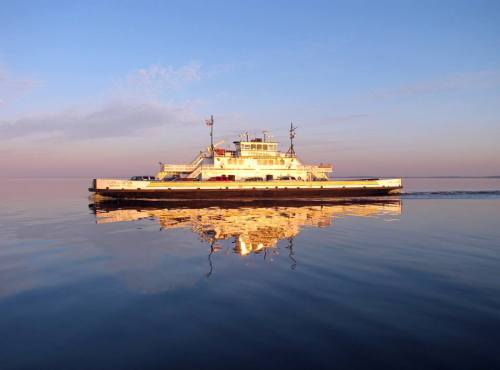 A ferry glides across calm waters at sunset, reflecting its image on the serene surface.