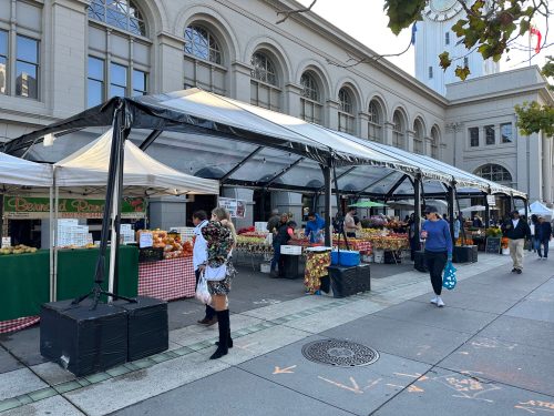 A bustling outdoor market with tents selling fresh produce, people browsing, and a historic building in the background.