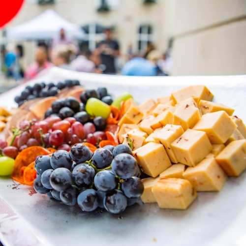 A colorful platter featuring assorted cheeses, grapes, and sliced fruits, set outdoors at a gathering.