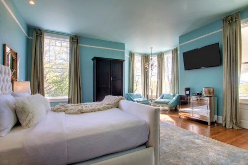 Bright, airy bedroom with a white bed, turquoise walls, large windows, and a cozy seating area.