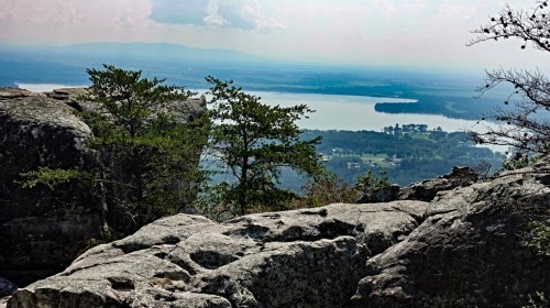 A scenic view from a rocky overlook, featuring a lake and distant mountains under a cloudy sky.