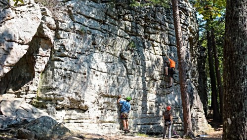 Two climbers ascend a rocky wall in a forest, while a third person observes from the ground.