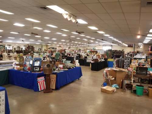 A spacious indoor market filled with various antique items, tables, and displays under bright overhead lights.
