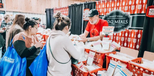 A vendor assists customers at a food booth, surrounded by baskets of meal packages in a lively market setting.