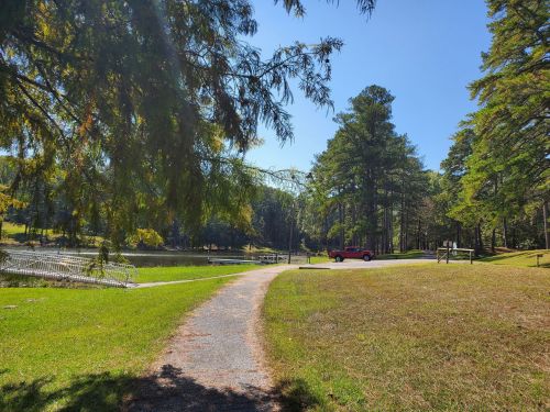 A sunny park scene with a path leading to a pond, surrounded by trees and a red vehicle parked nearby.