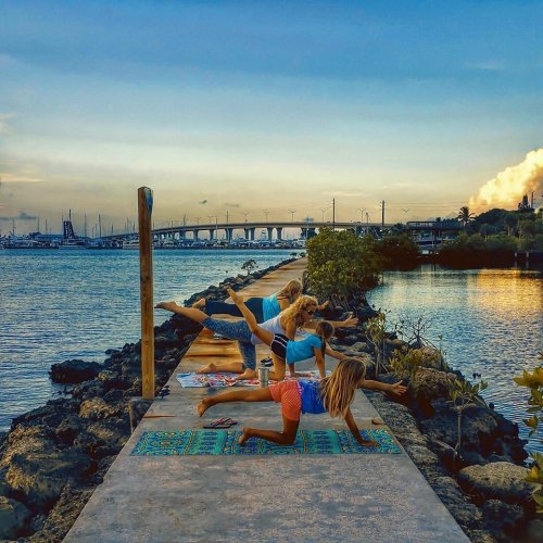 Three women practicing yoga on a pier at sunset, with a scenic view of water and a bridge in the background.