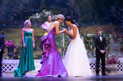 Two women in elegant gowns share a joyful moment on stage, surrounded by floral decorations and an audience.