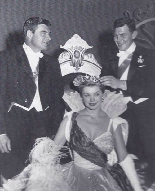 A woman in a gown and crown is seated, while two men in formal attire place a tiara on her head.