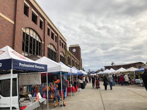 A bustling outdoor market with vendor tents, shoppers, and a brick building in the background under a cloudy sky.