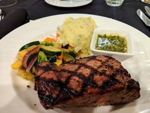 A plate featuring grilled steak, mashed potatoes, sautéed vegetables, and a small dish of green sauce.
